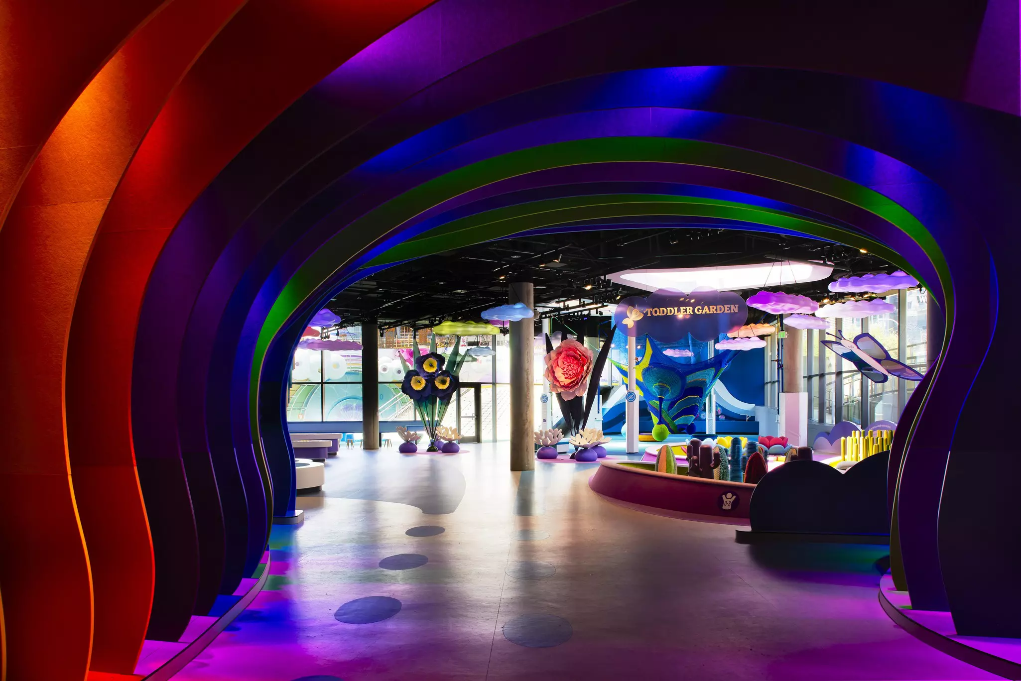 Brightly colored curved entryway to a children's museum with "Toddler Garden" on a sign in the distance, along with tall, oversized art of butterflies and flowers.