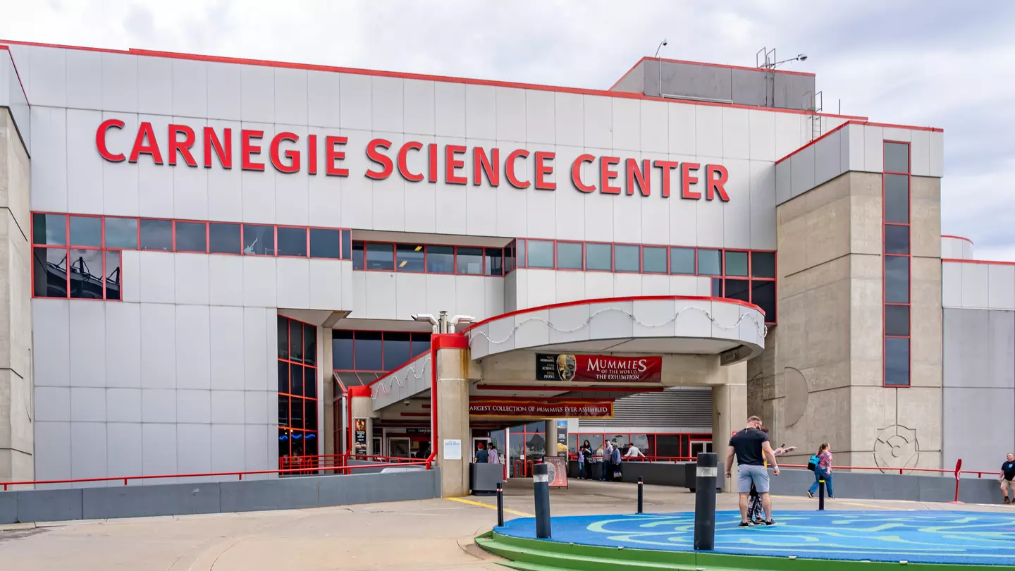 Entrance of Carnegie Science Center in Pittsburgh, Pennsylvania There are a few people standing outside of the red and gray building