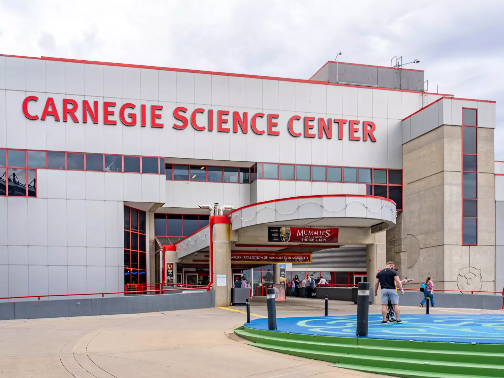 Entrance of Carnegie Science Center in Pittsburgh, Pennsylvania There are a few people standing outside of the red and gray building