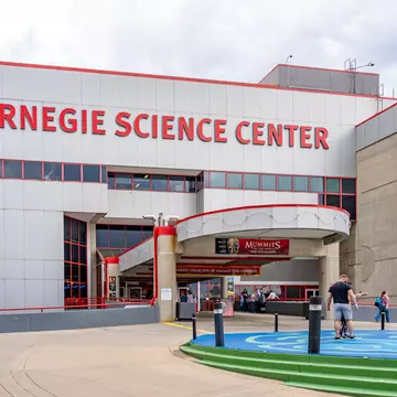 Entrance of Carnegie Science Center in Pittsburgh, Pennsylvania There are a few people standing outside of the red and gray building