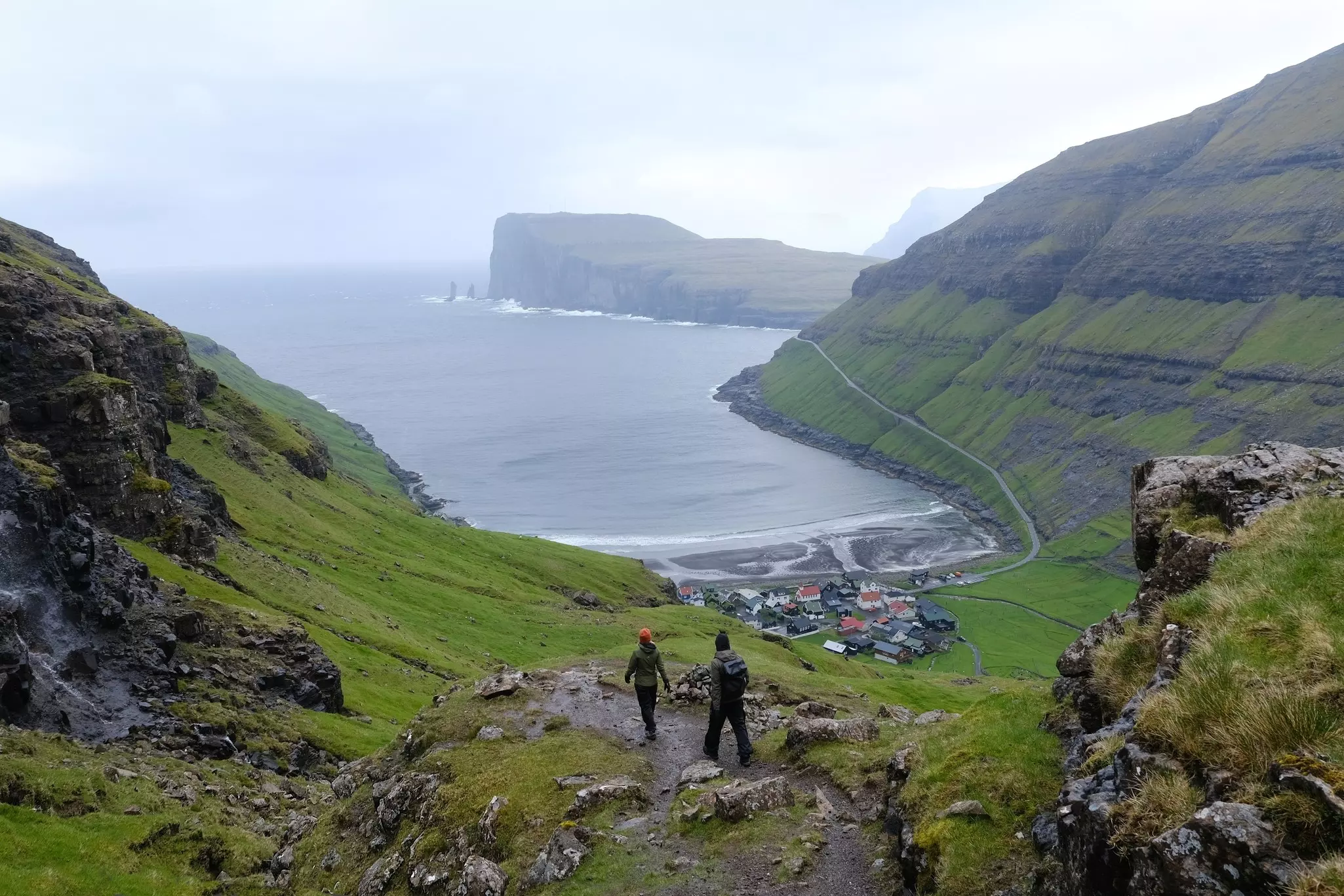 Two hikers stand at a promontory looking down at a cove surrounded by steep mounatins. The surf hits rock formations in the distance,