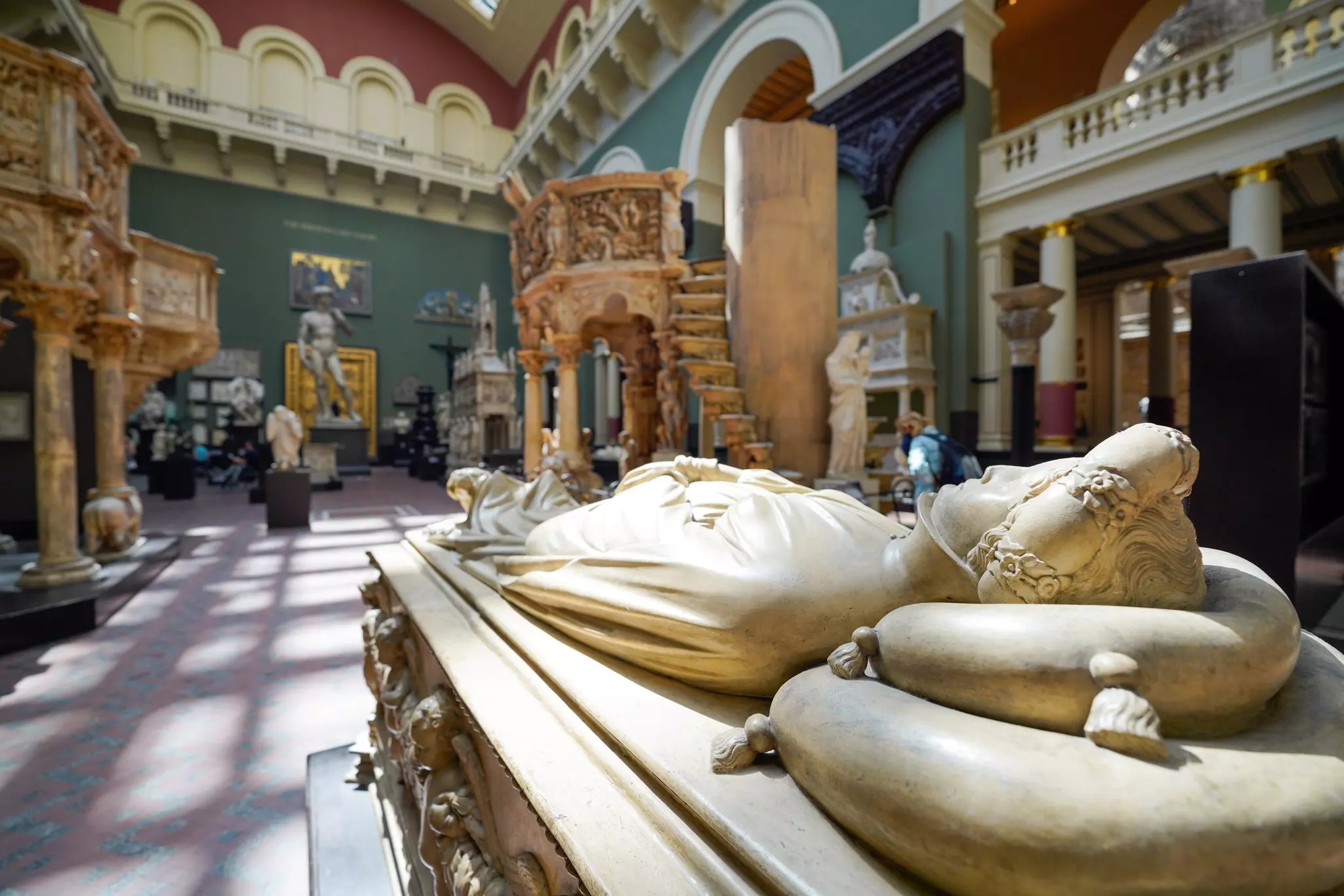 Marble sculpture of a human figure lying prone with other statues and ornate surroundings inside a bright museum with sunlight streaming in from ceiling windows.