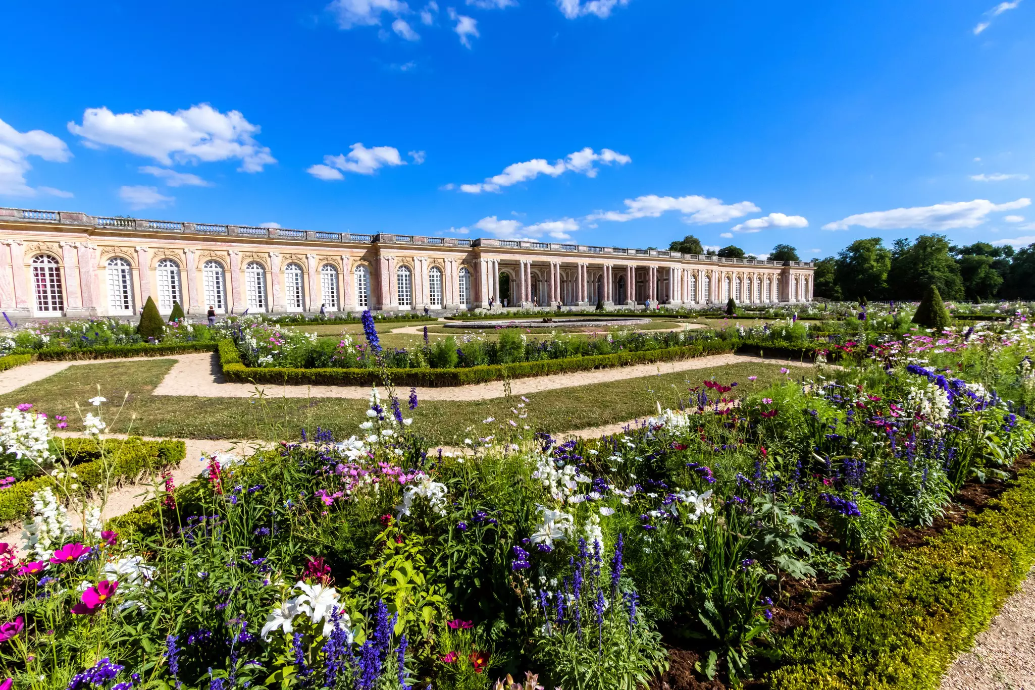 Flowers and hedges around paths with a long building with arched windows in the background.