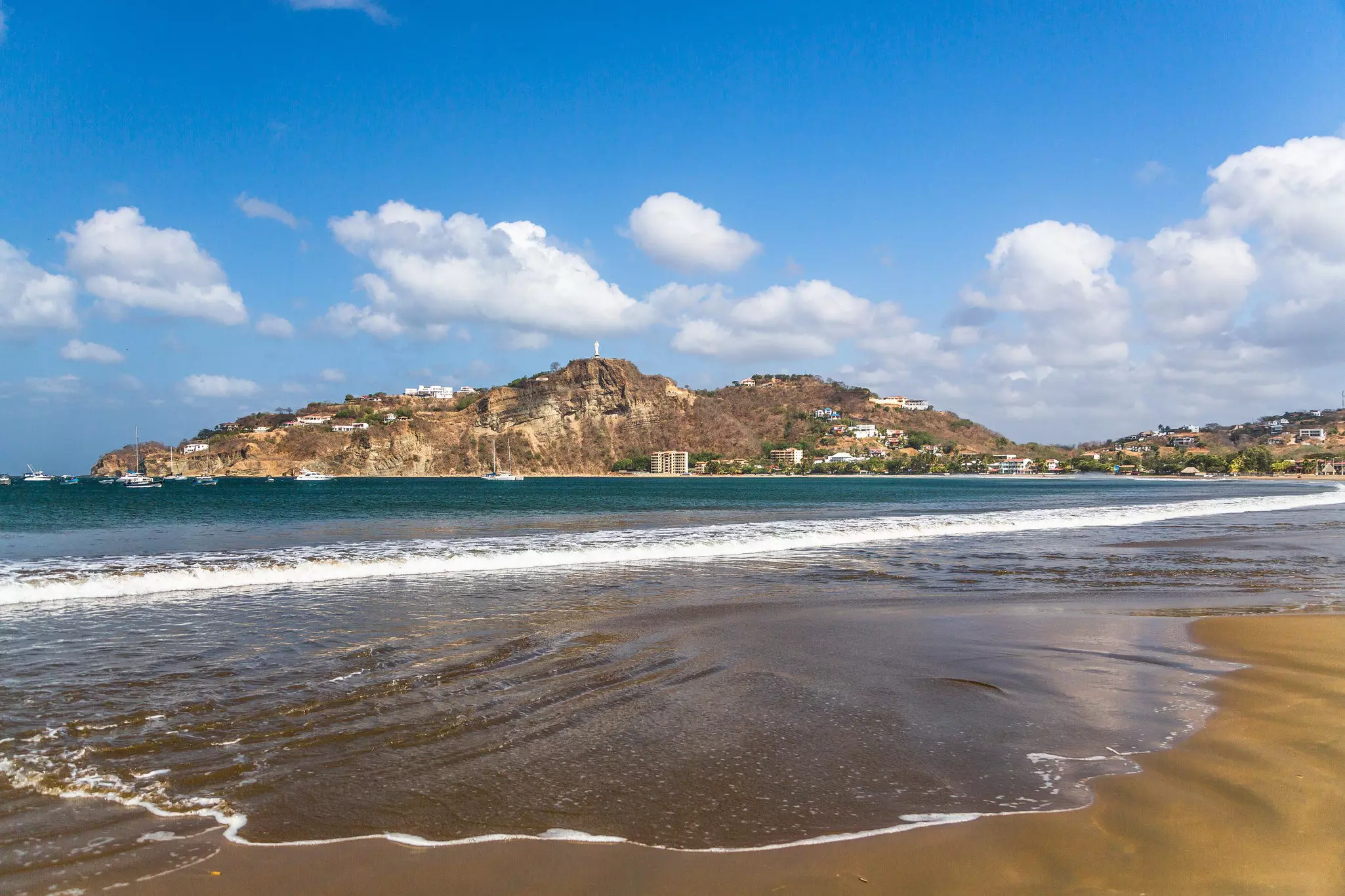 Gentle waves lap the sandy light brown shores. In the background there's a large mountain dotted with homes and hotels. In the distance, a few boats are moored in the ocean.