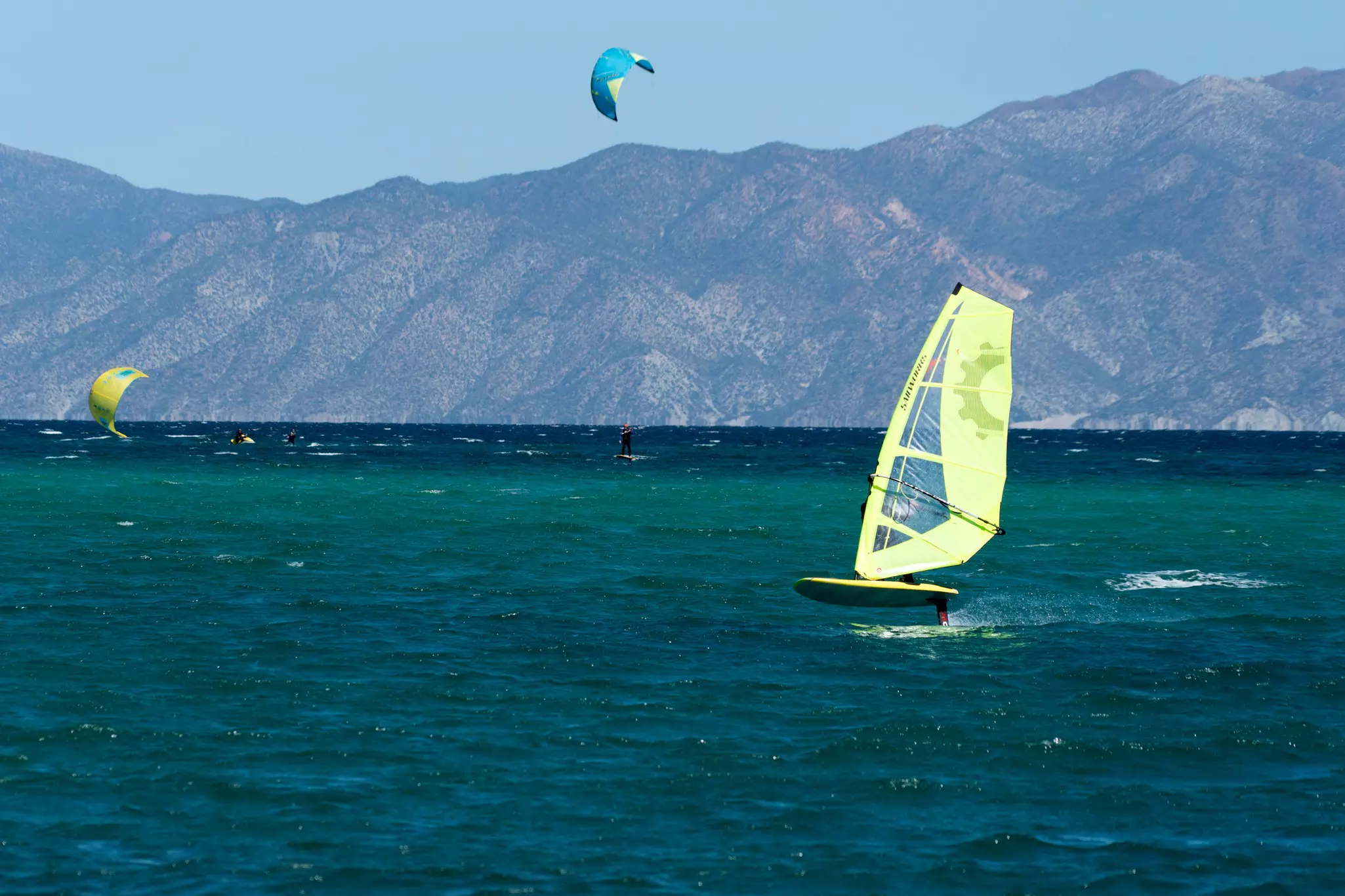 Sails of windsurfers and kiteboarders are seen on the water. Mountains rise from the coast in the distance.