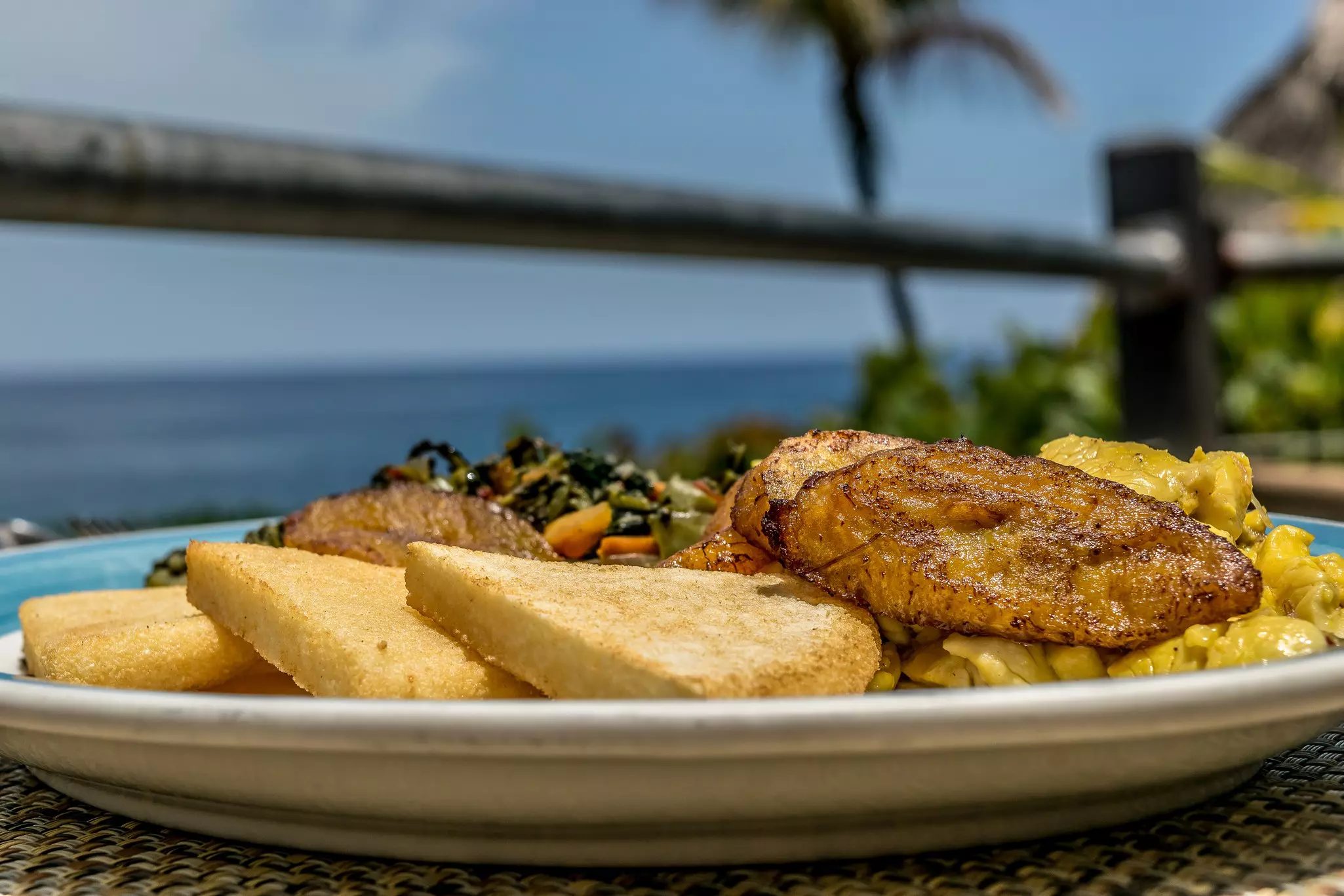 A plate at an outside table near the sea loaded with fried bread, plantain and saltfish.