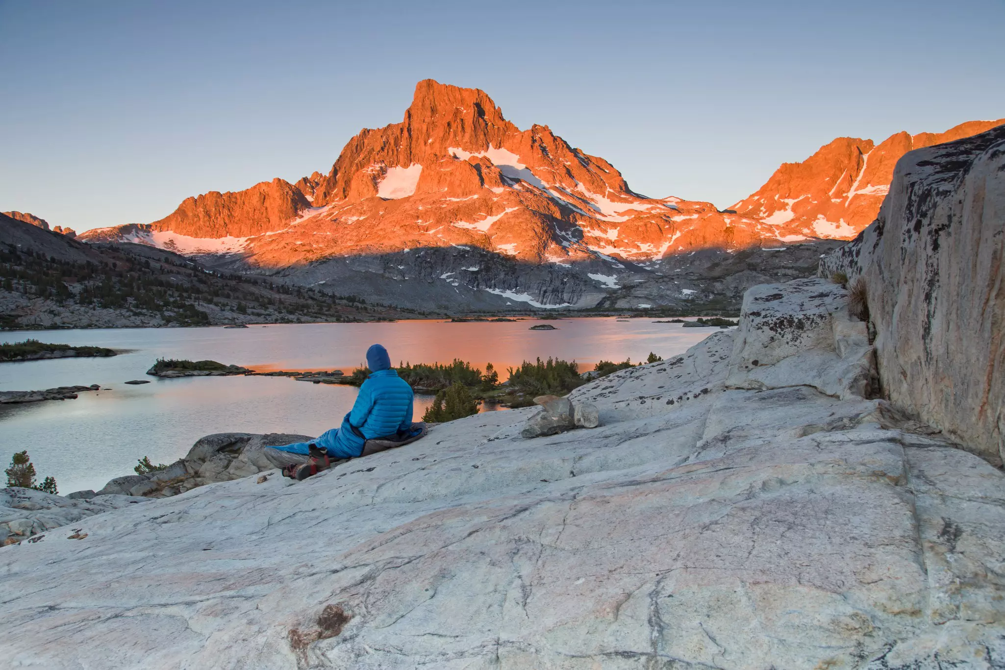A male hiker reclines on a rock as he watches the sunrise on a distant mountain.