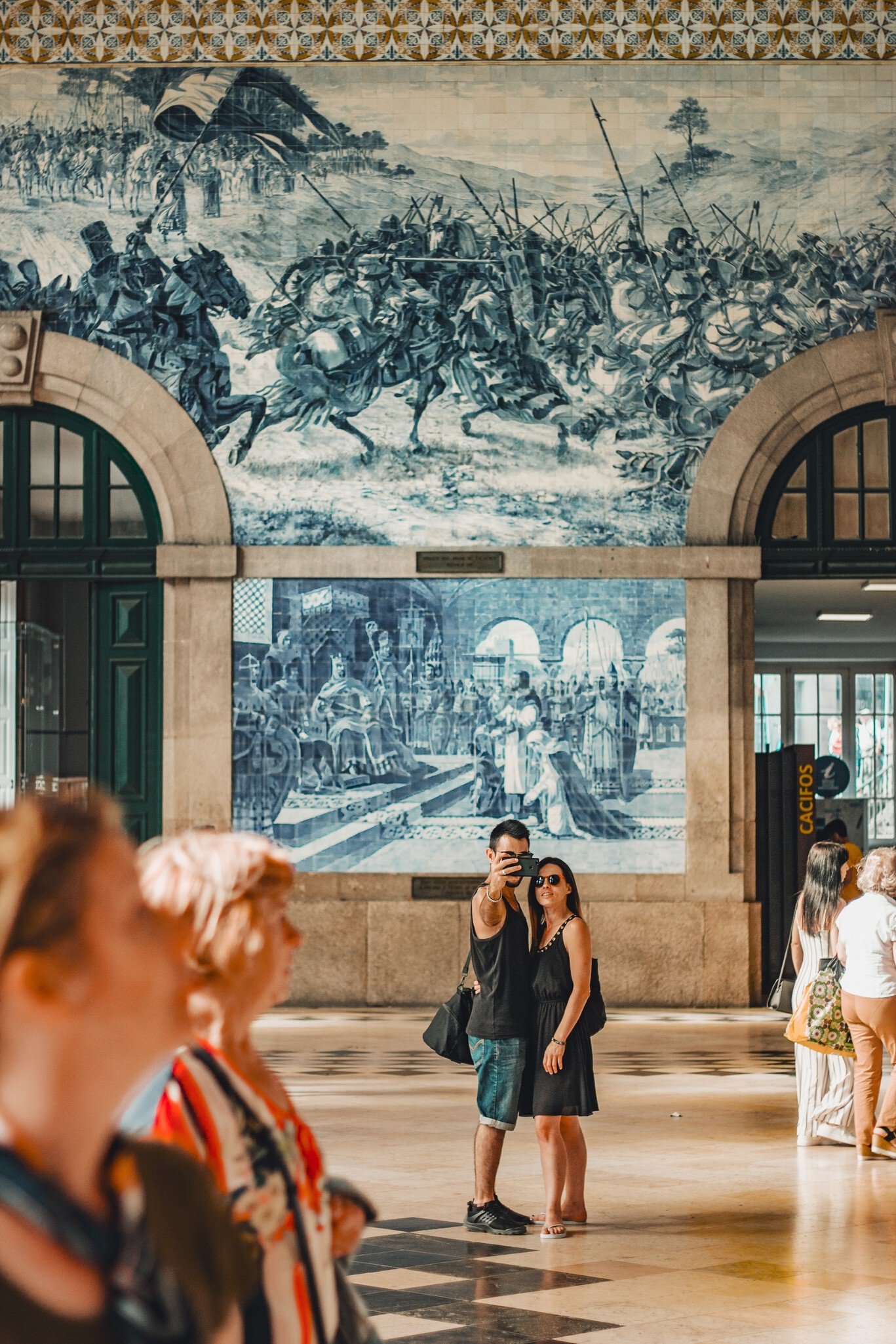 A couple taking a selfie in front of a traditional and typical blue tile art in São Bento train station in Porto, Portugal.