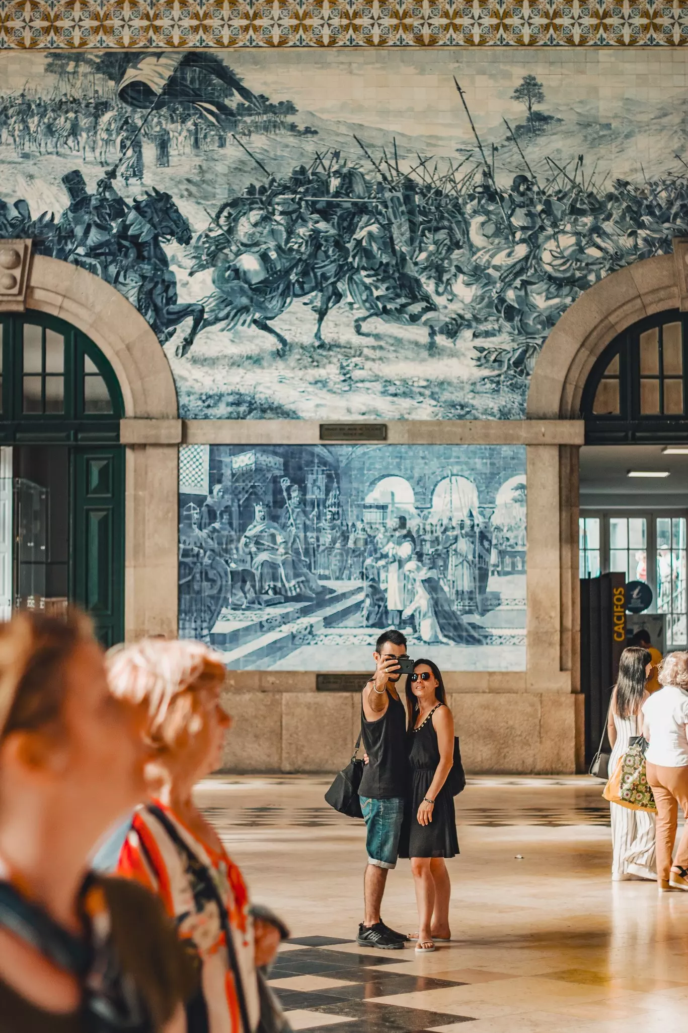 Couple of tourist taking a selfie in front of a traditional and typical blue tile art in São Bento train station in Oporto, Portugal in a hot sunny day.