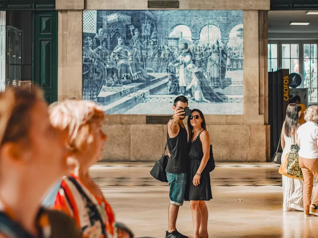 A couple taking a selfie in front of a traditional and typical blue tile art in São Bento train station in Porto, Portugal.