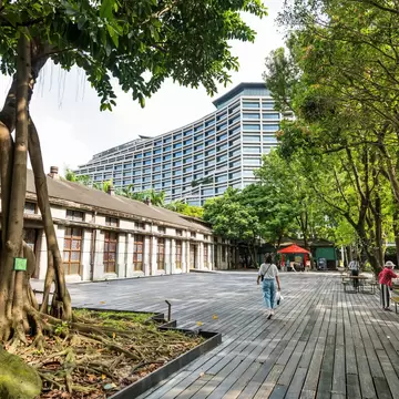 Wooden deck with picnic tables beside long low heritage factory buildings, big banyan trees and a modern tower behind