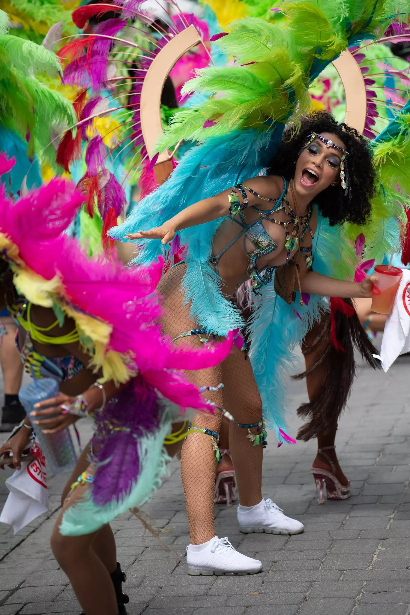 A woman in a colorful feathered costume holding a cup with a straw and dancing with others similarly dressed