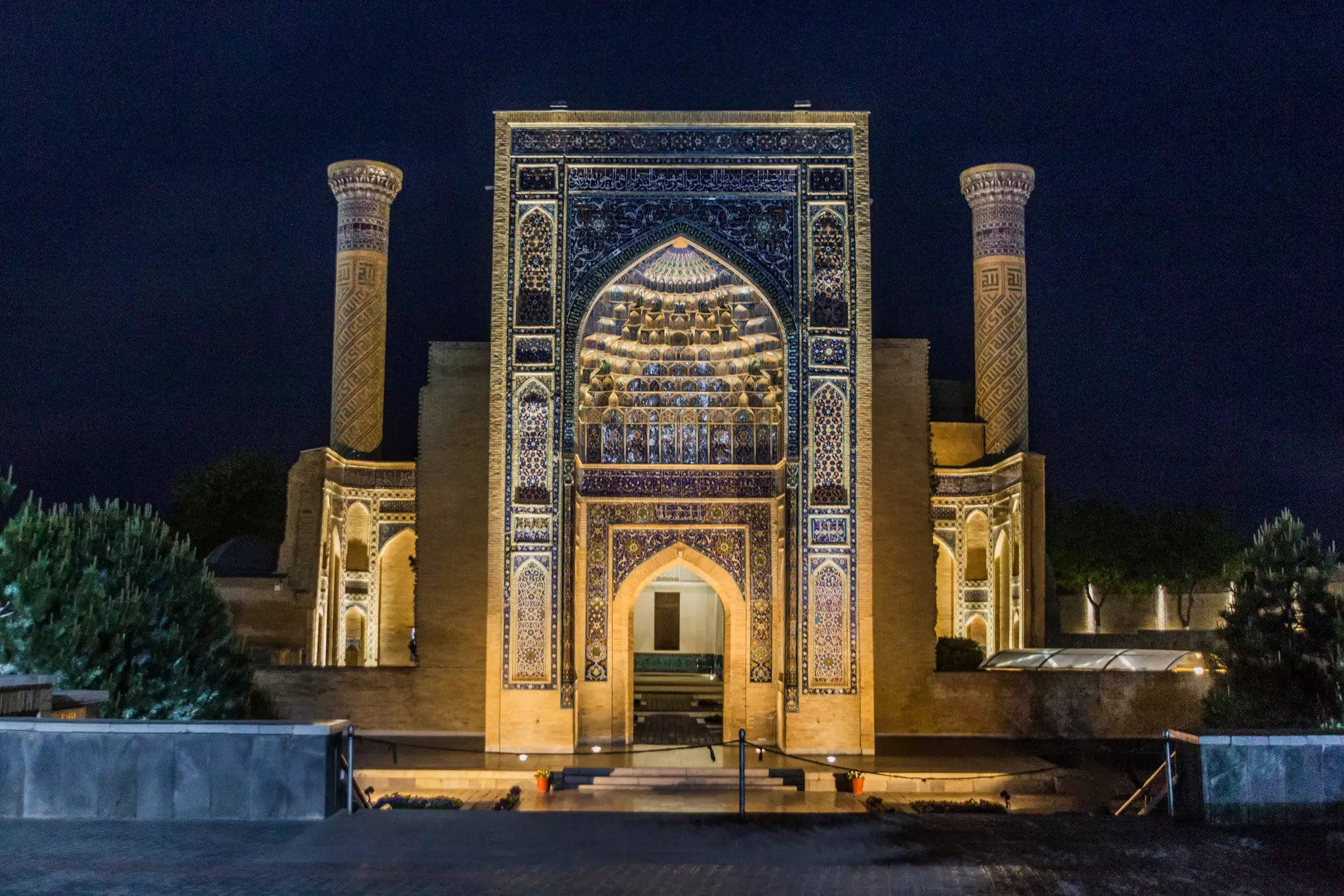 An evening view of the illuminated facade of a mausoleum, with blue tile in niches, Islamic arches and other details.