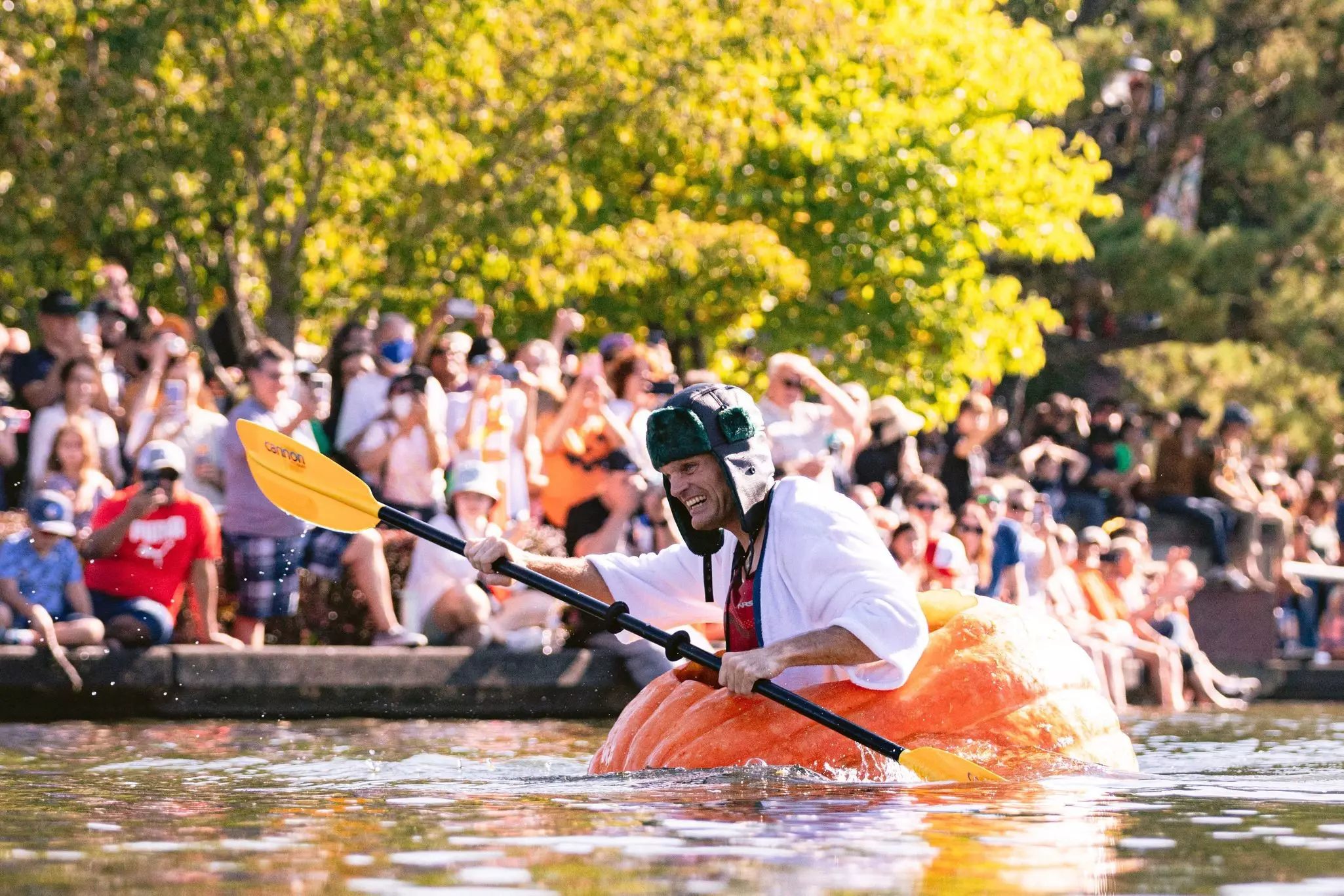 A man in a winter hat paddles in a giant pumpkin in water as crowds look on on a sunny fall day