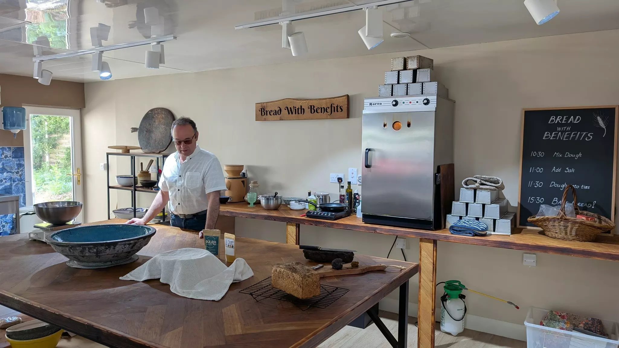 A man stands at a kitchen counter in front of a sign that says "bread with benefits"