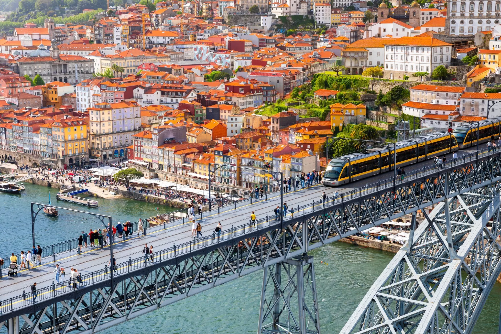 Aerial view of bridge with pedestrians and tram with city of red-roofed buildings in the distance.