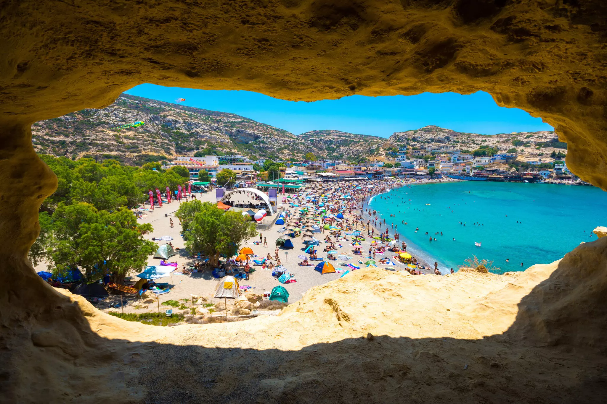 A view of a crowded beach with colorful umbrellas and a village beyond from a cave overlooking the scene.