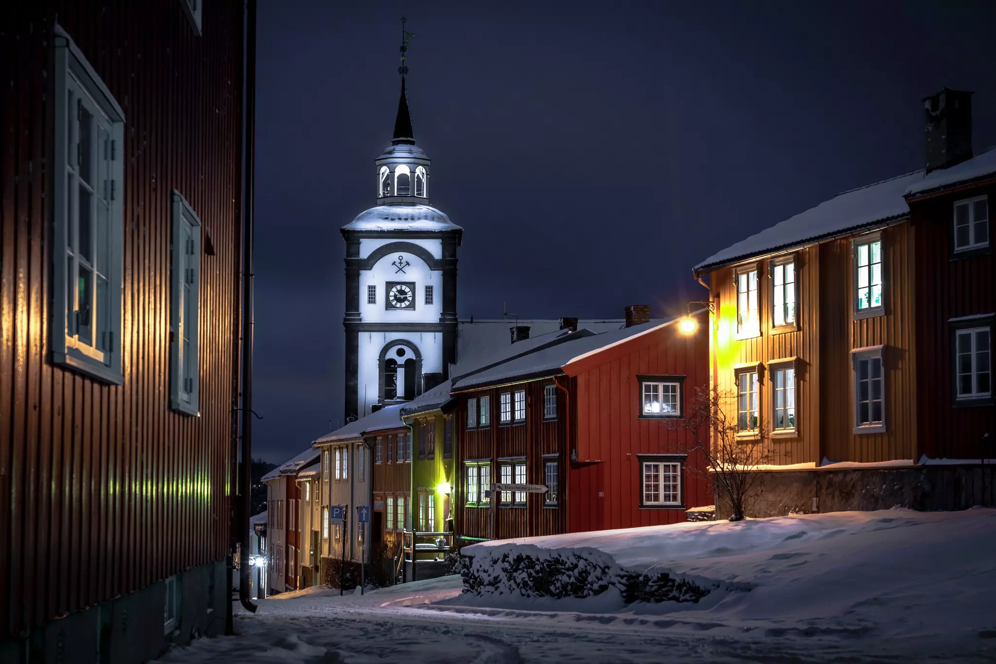 The white tower of a church is seen at night in a historic village. Snow blankets the ground.