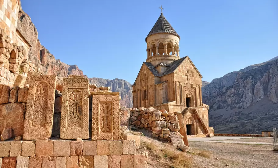 Noravank Monastery in Armenia: two-storey stone church with a drum belfry, carved cross-stones in front, red cliffs behind