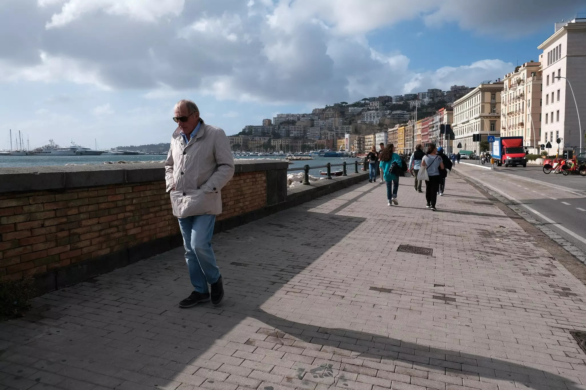 A senior man wears a coat as he walks along a waterfront sidewalk. Apartment buildings lining the street by the shore are seen behind him.
