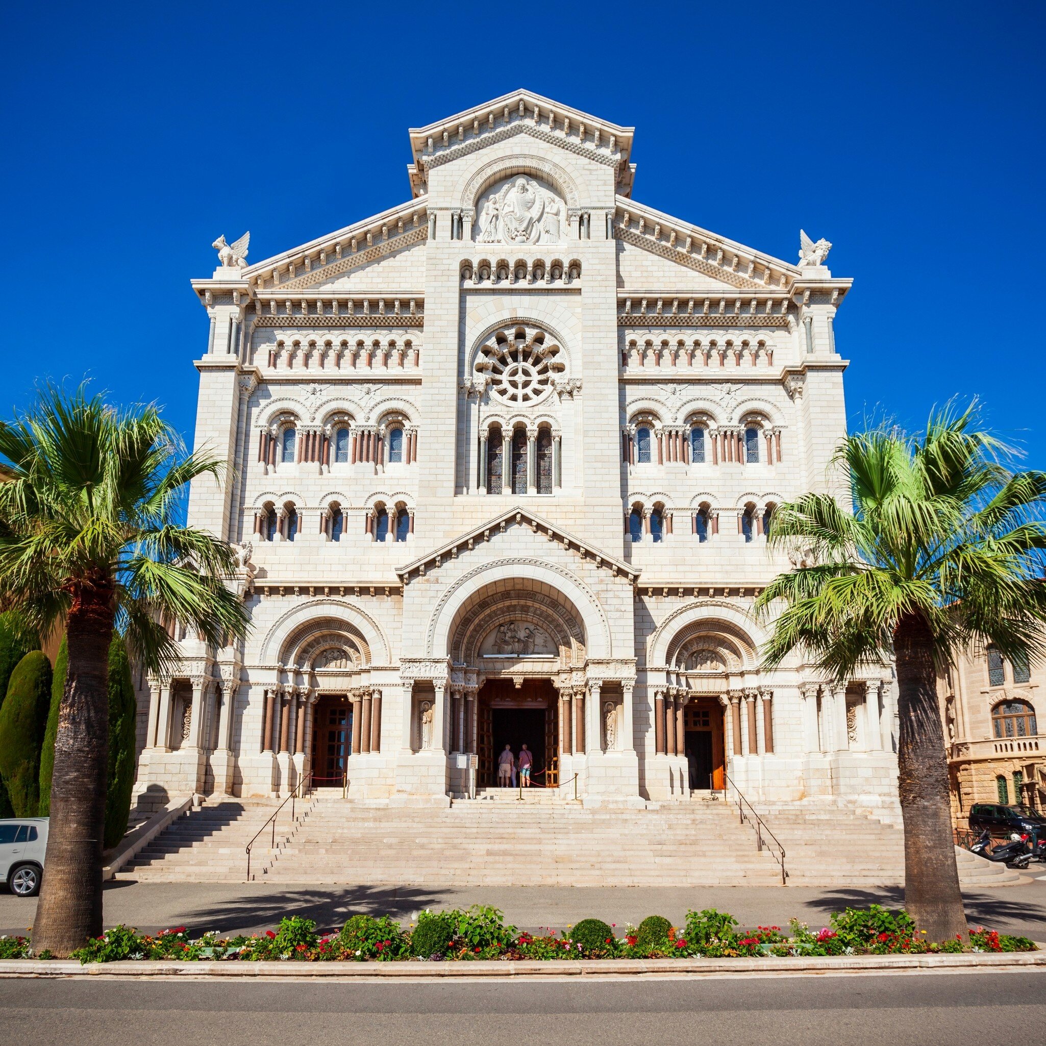 White stone church front with a rose window and three arched doorways above wide steps, framed by two palm trees