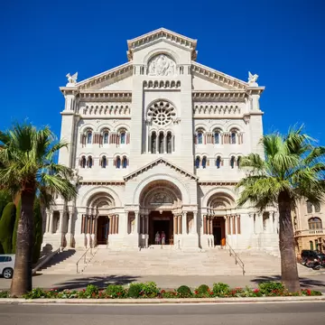 White stone church front with a rose window and three arched doorways above wide steps, framed by two palm trees
