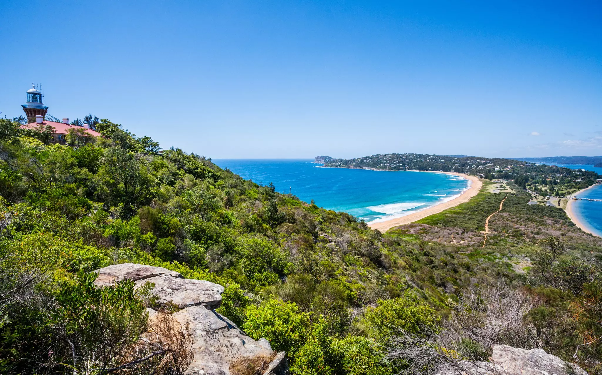 A lighthouse tops a hillside above a curve of beach on a spit of land with ocean each side.