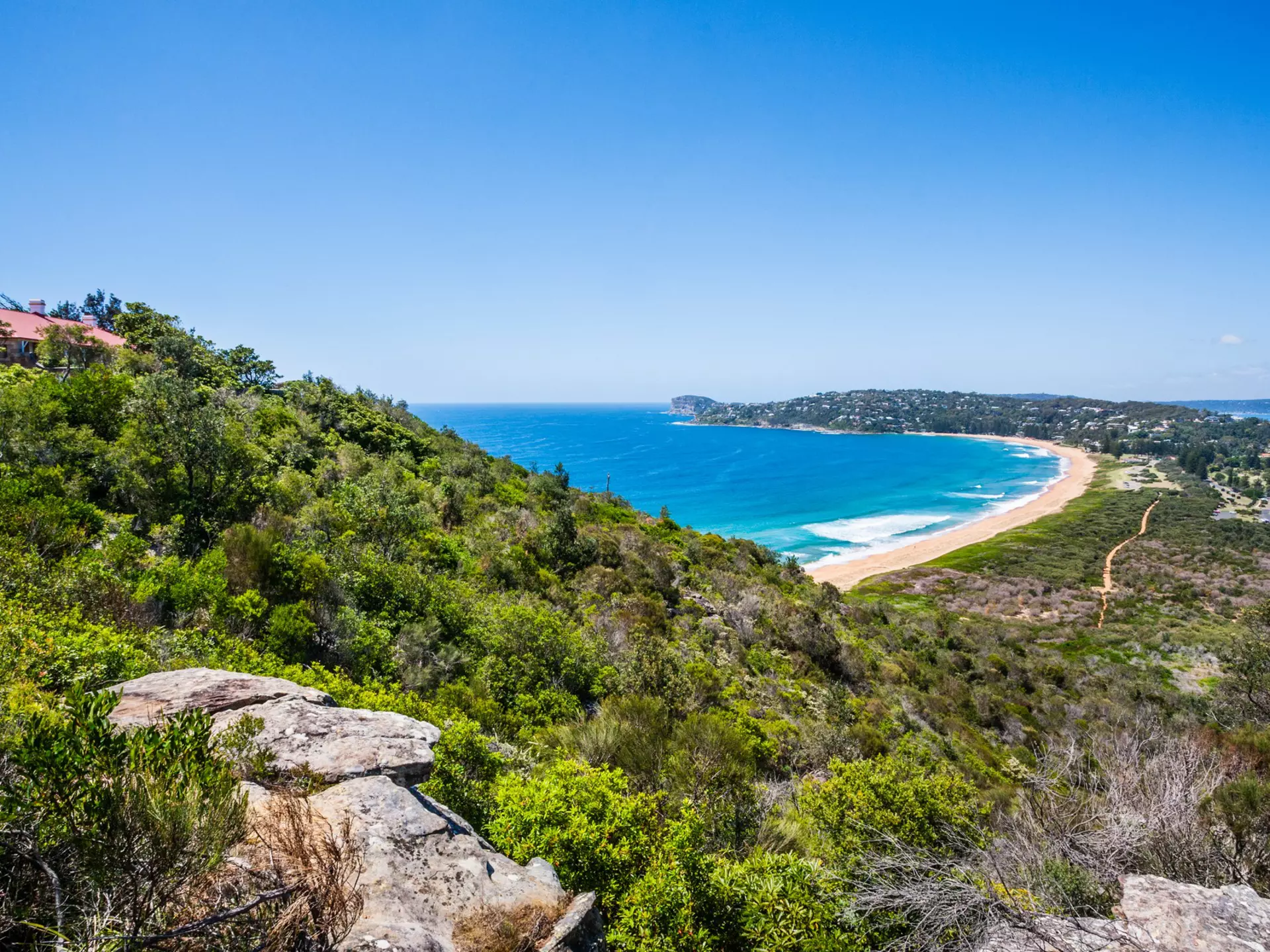 A lighthouse tops a hillside above a curve of beach on a spit of land with ocean each side.