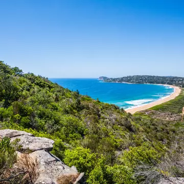 A lighthouse tops a hillside above a curve of beach on a spit of land with ocean each side.