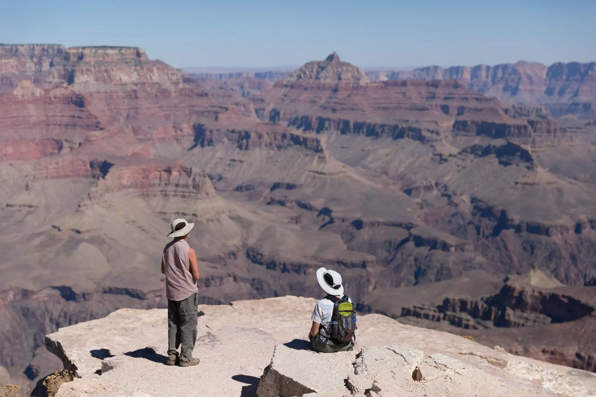 One person standing and another sitting, contemplating the wide landscape of Grand Canyon at Shoshone Point.