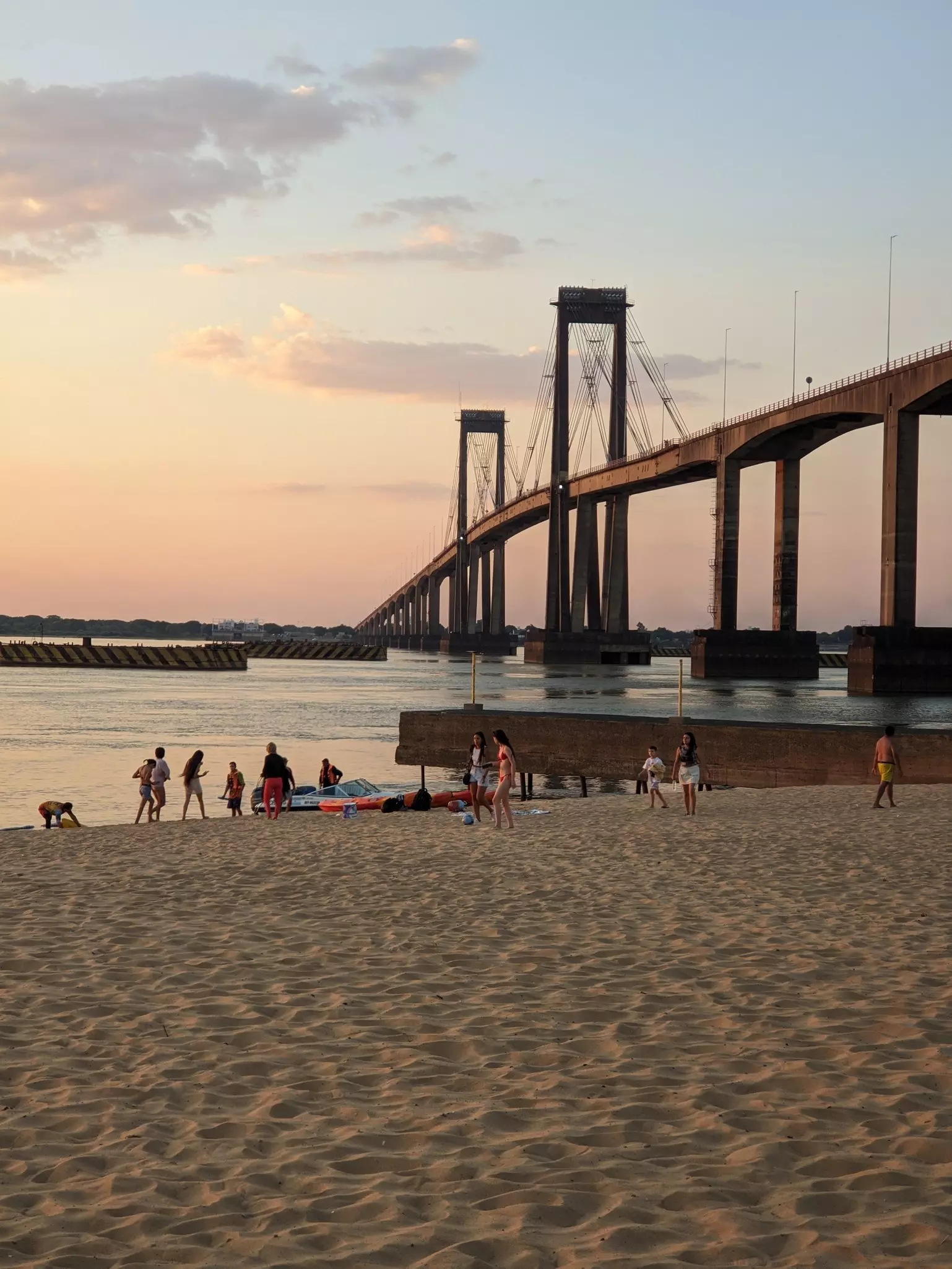 People on a riverside beach in the shadow of a large suspension bridge in the evening light.