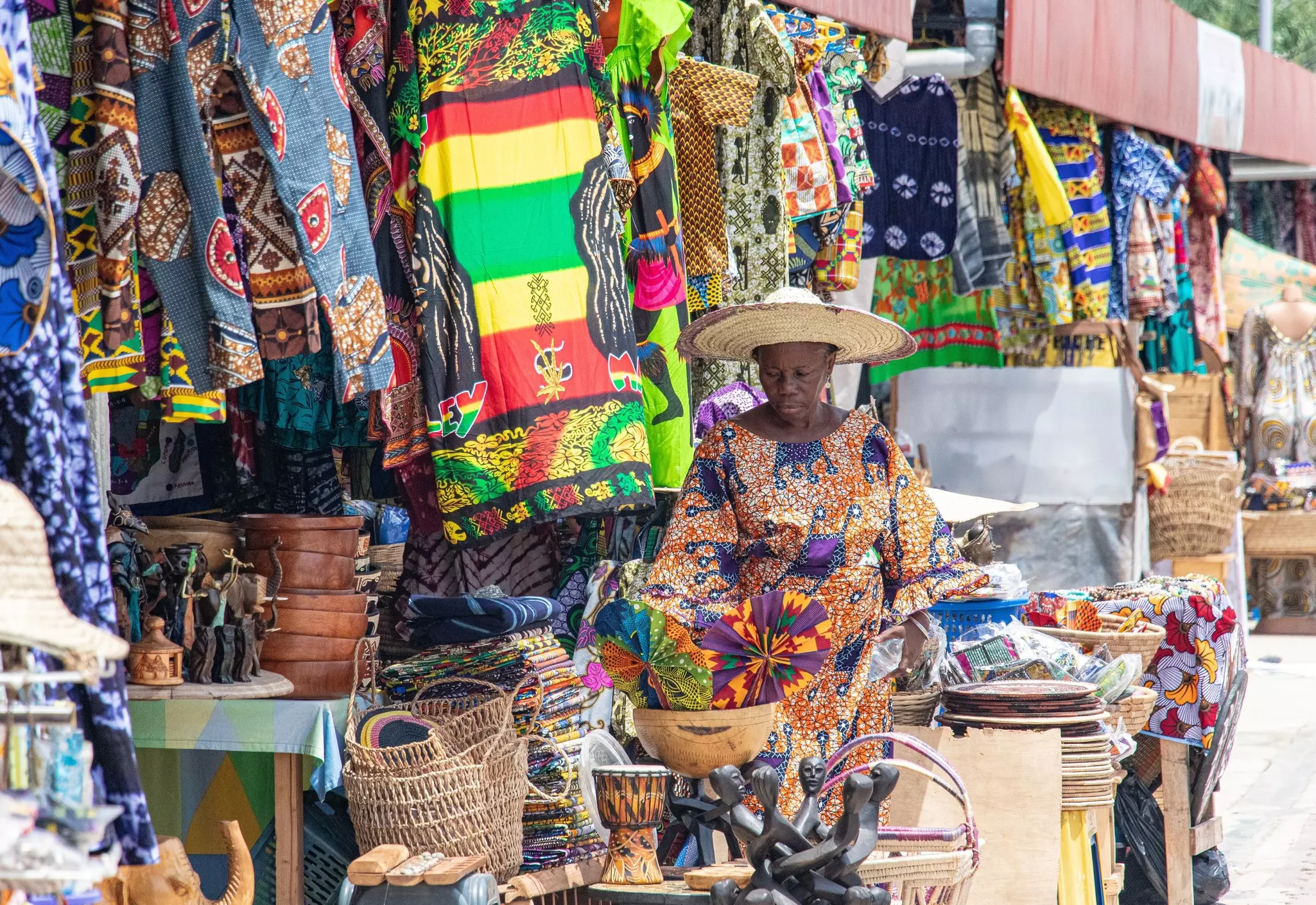 A woman in a straw hat sits at a market stall filled with baskets and textiles for sale.