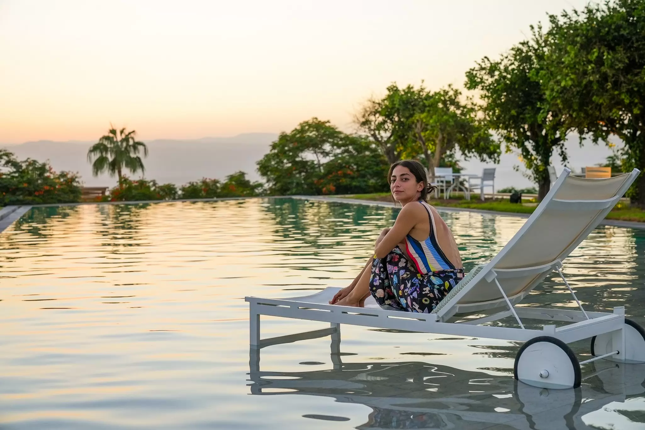 Woman on a floating lounger in a calm infinity pool overlooking distant hills at sunset