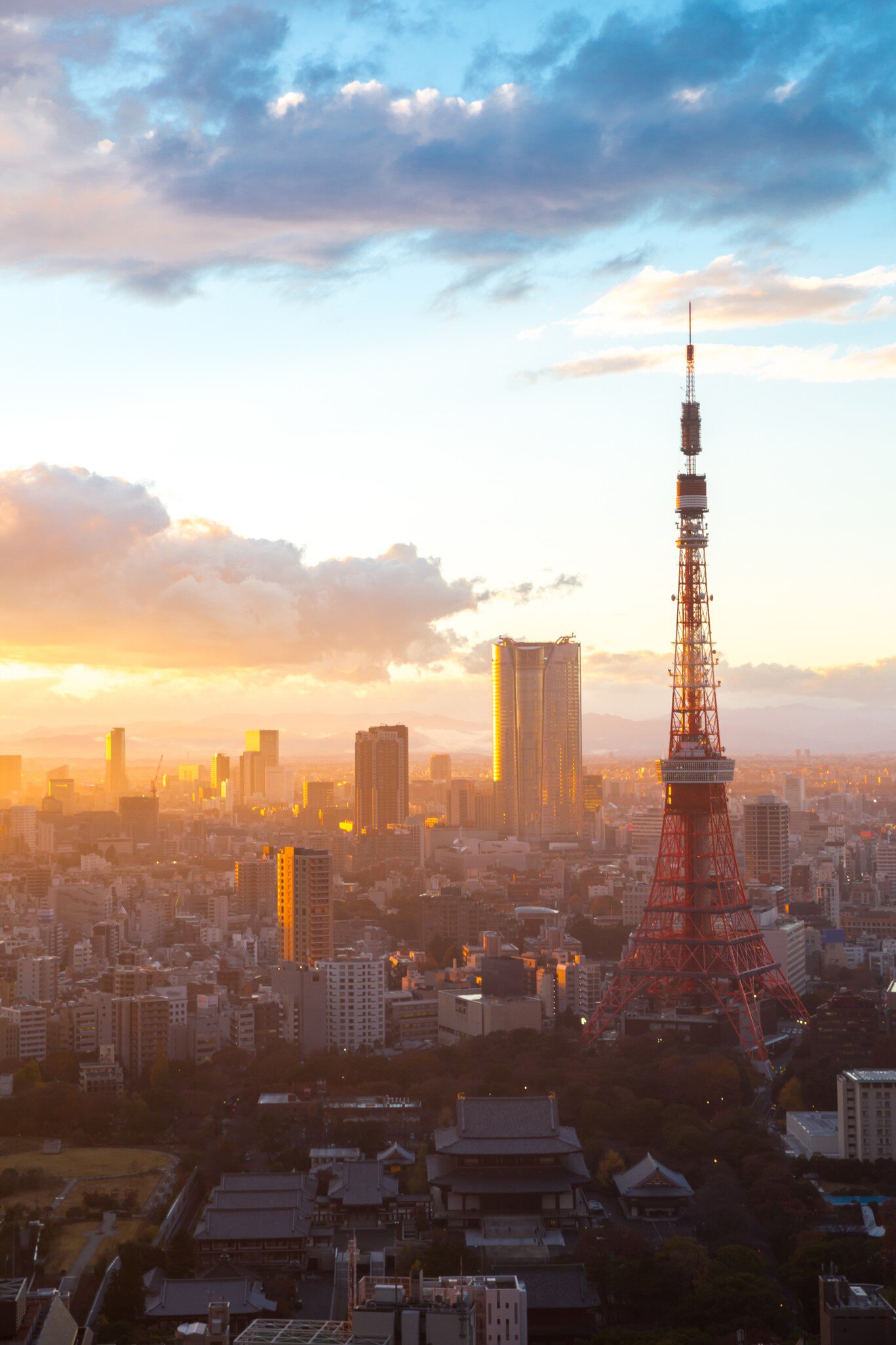 Red and white Tokyo Tower beside dark temple roofs at sunset, with city towers glowing in golden light under clouds.