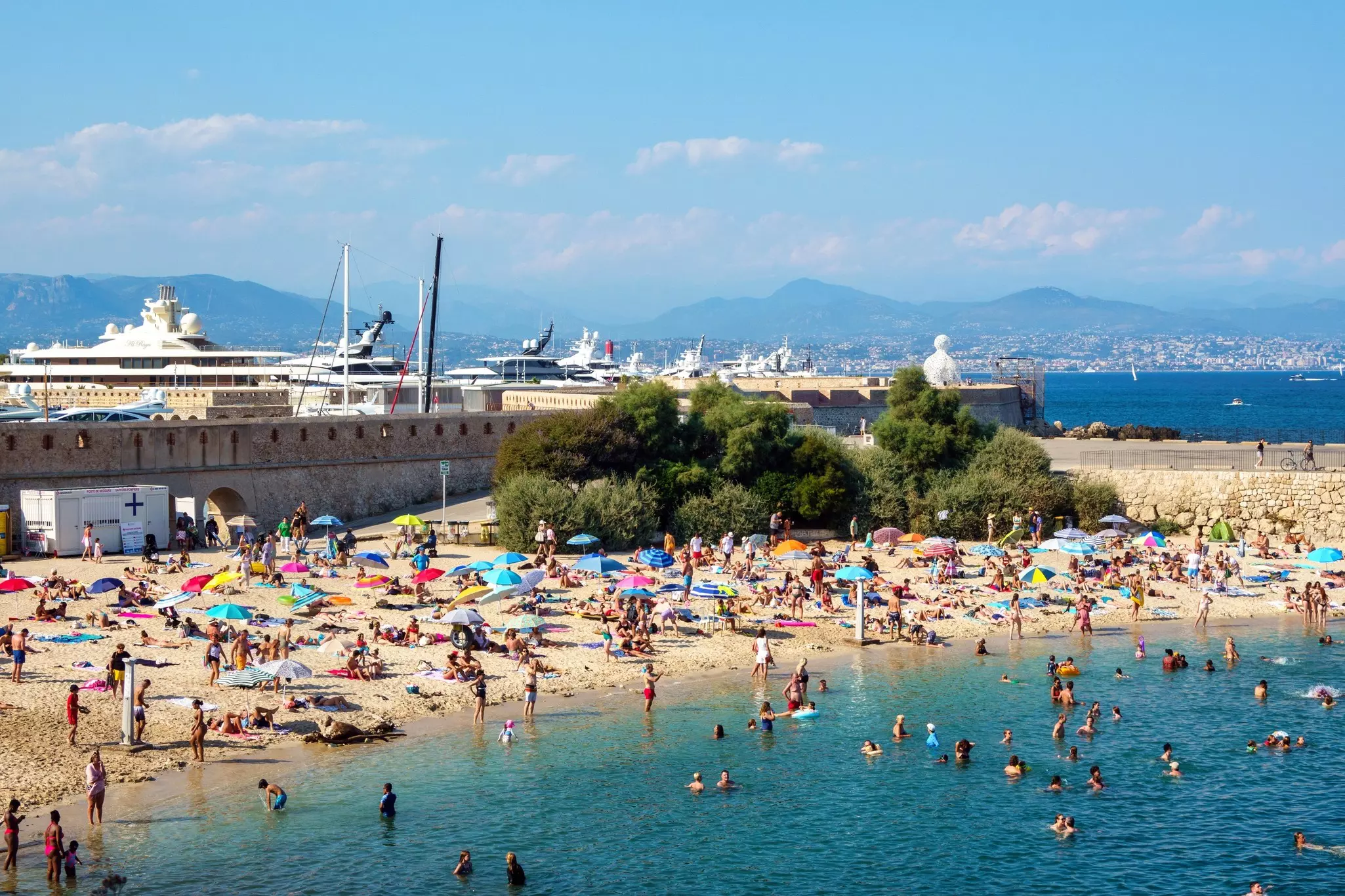 People swim off a sandy bay. A large sculpture sits on the harbor wall behind where several yachts are docked.