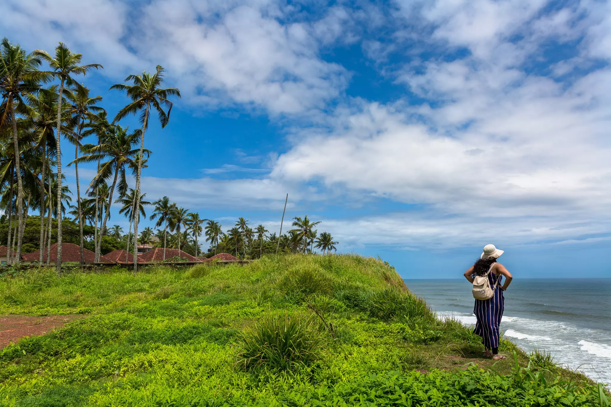 A solo female traveler stands on a cliff looking out to sea in a rural area with palm trees and small houses.