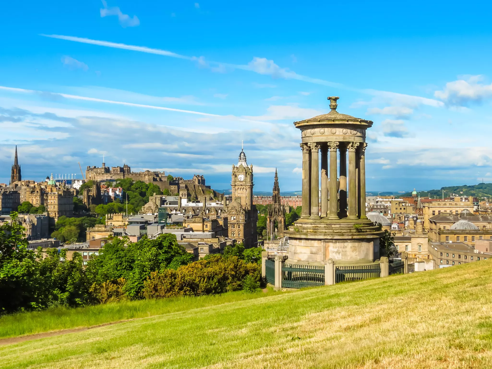 Round, columned monument amid grassy field with historic city skyline in the distance on a sunny day.