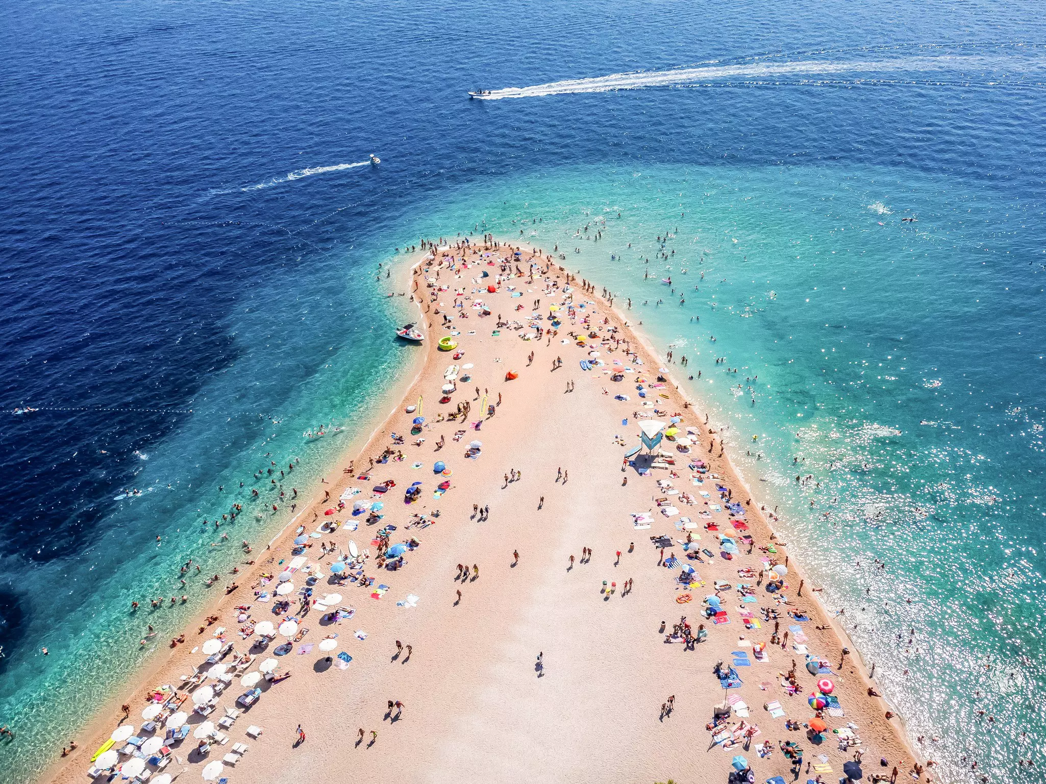 An aerial view of a beach forming a point, with hundreds of sunbathers and swimmers.