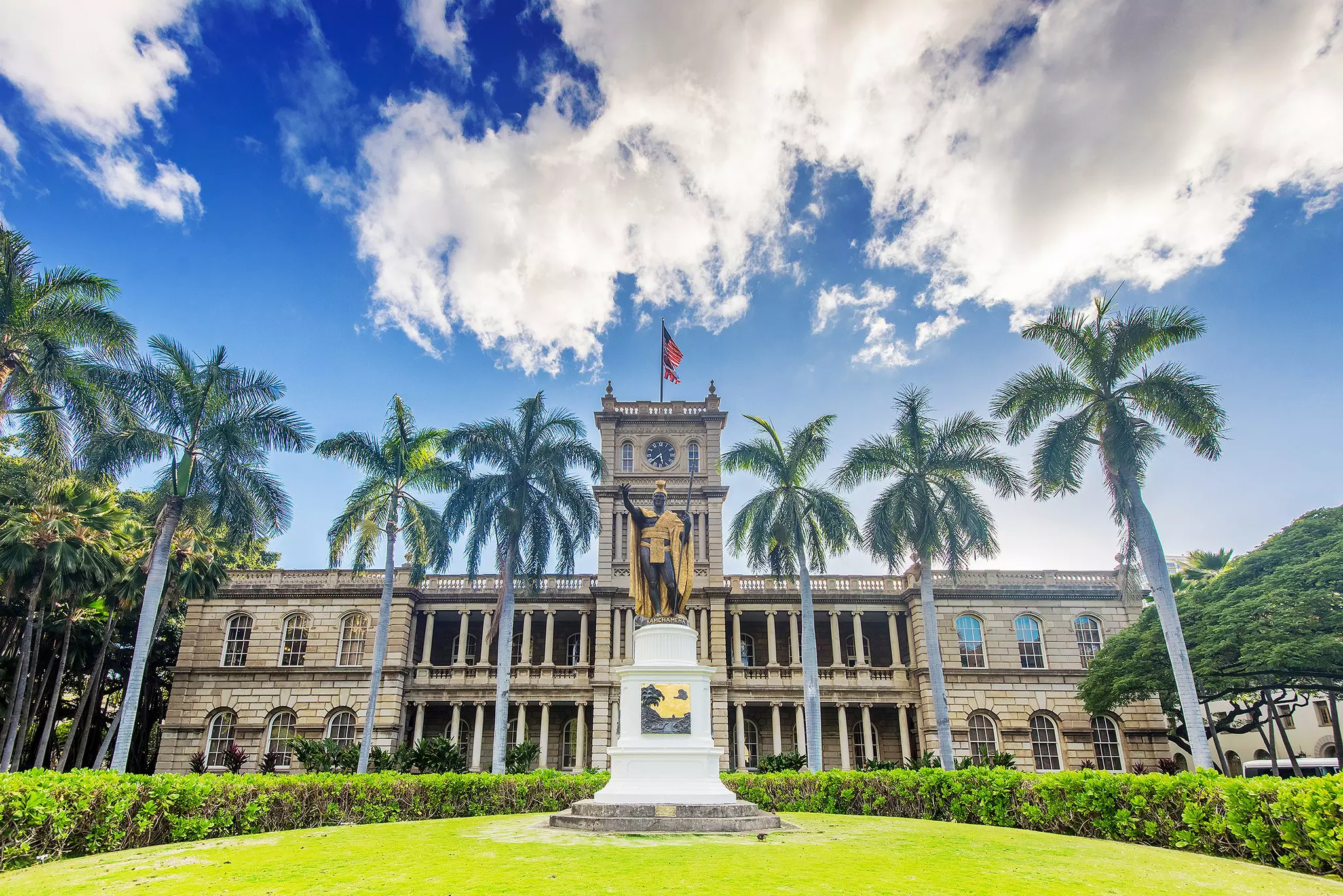 Bronze statue of man holding a staff situated on a grassy knoll ringed by palm trees with a large stone palace in the background on a sunny day.