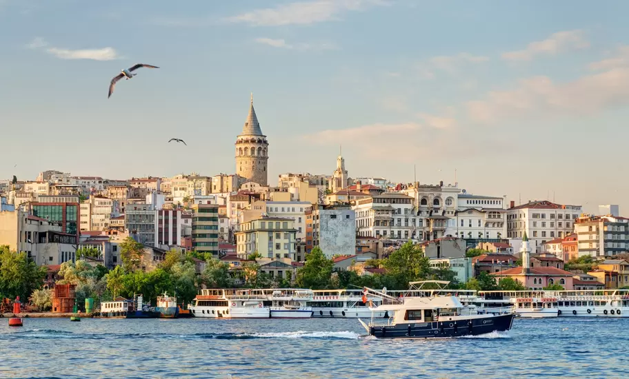 Galata Tower rising above Beyoğlu's hillside buildings in İstanbul, with ferries moored and a motor yacht on the Golden Horn