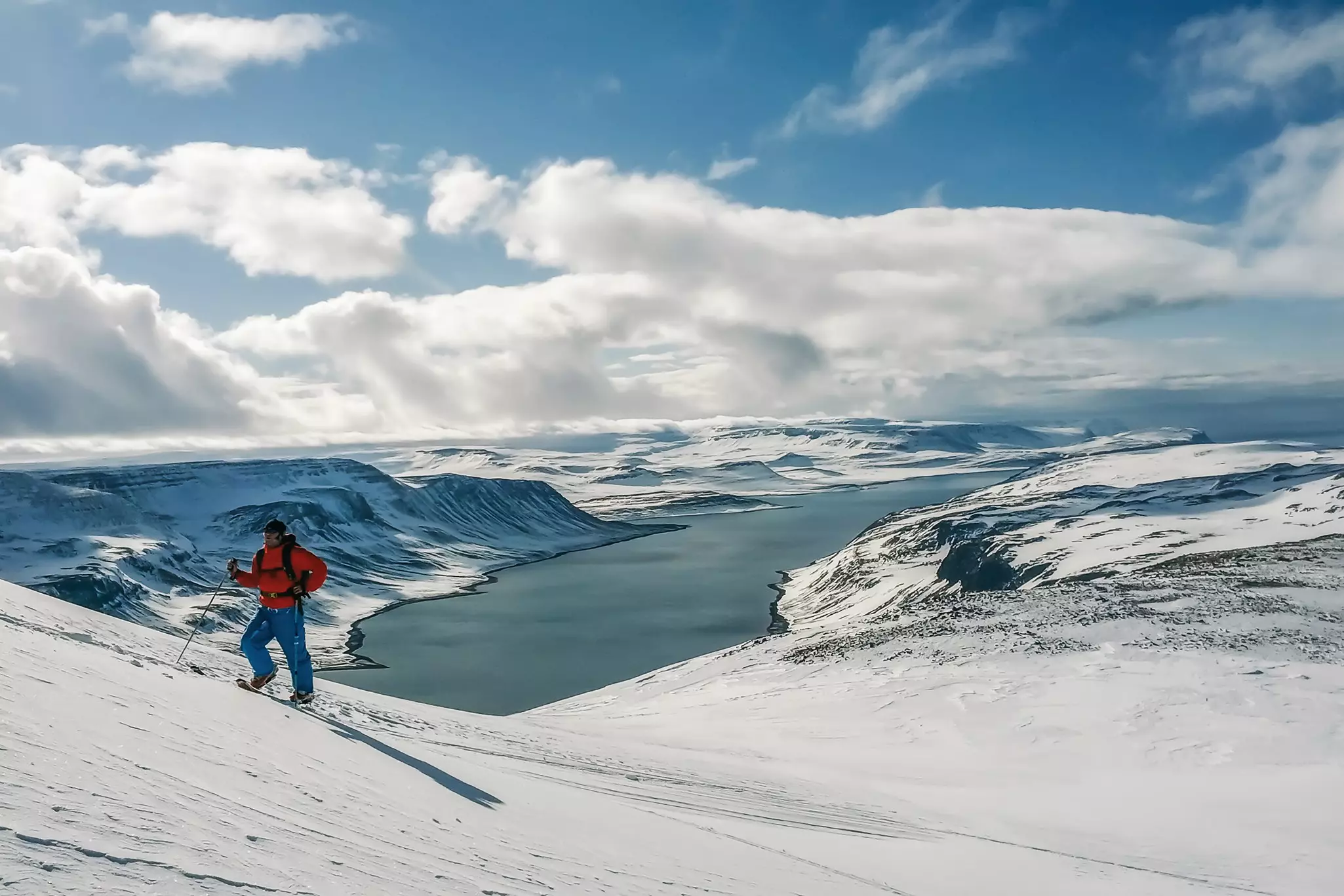 A mountaineer in red snow gear hikes up a snow-covered hill above a fjord.