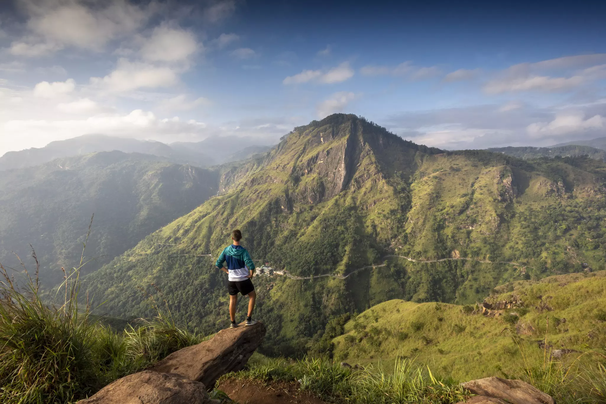 A man stands at a viewpoint on Little Adam's Peak in Sri Lanka, looking out across a hilly rural landscape.