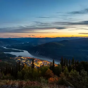 Dusk view over the lit-up town of Dawson City and the winding Yukon River from the forested summit of the Midnight Dome