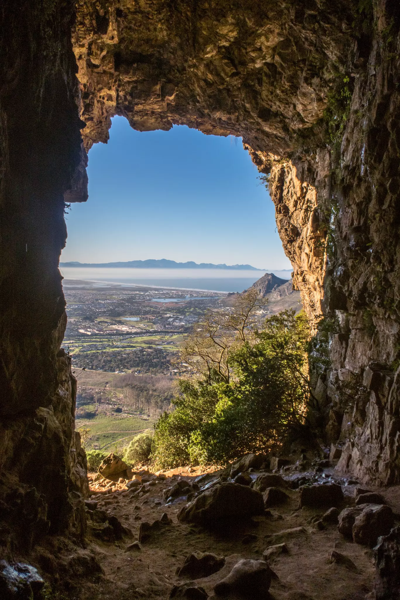 A view through a cave opening of a grassy landscape, mountains and buildings