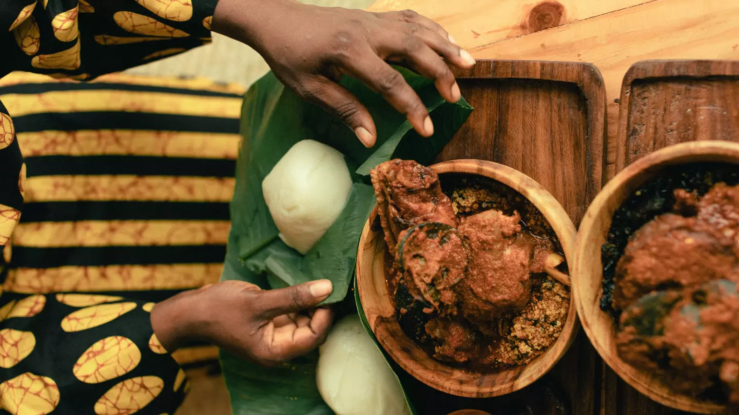 A woman’s hands are pictured from above as she unwraps leaf-covered yams. Two bowls of stew sit on the table next to the yams.