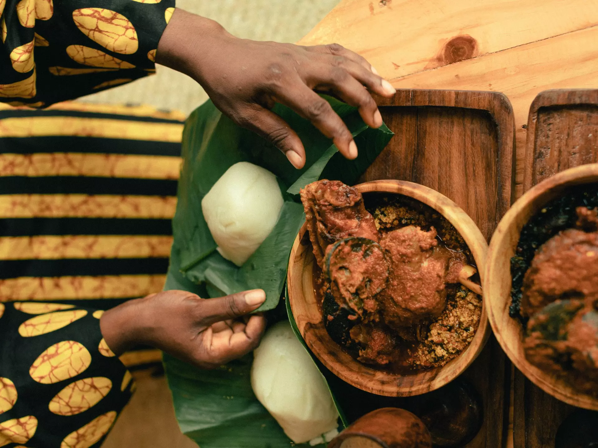 A woman’s hands are pictured from above as she unwraps leaf-covered yams. Two bowls of stew sit on the table next to the yams.