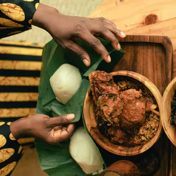 A woman’s hands are pictured from above as she unwraps leaf-covered yams. Two bowls of stew sit on the table next to the yams.
