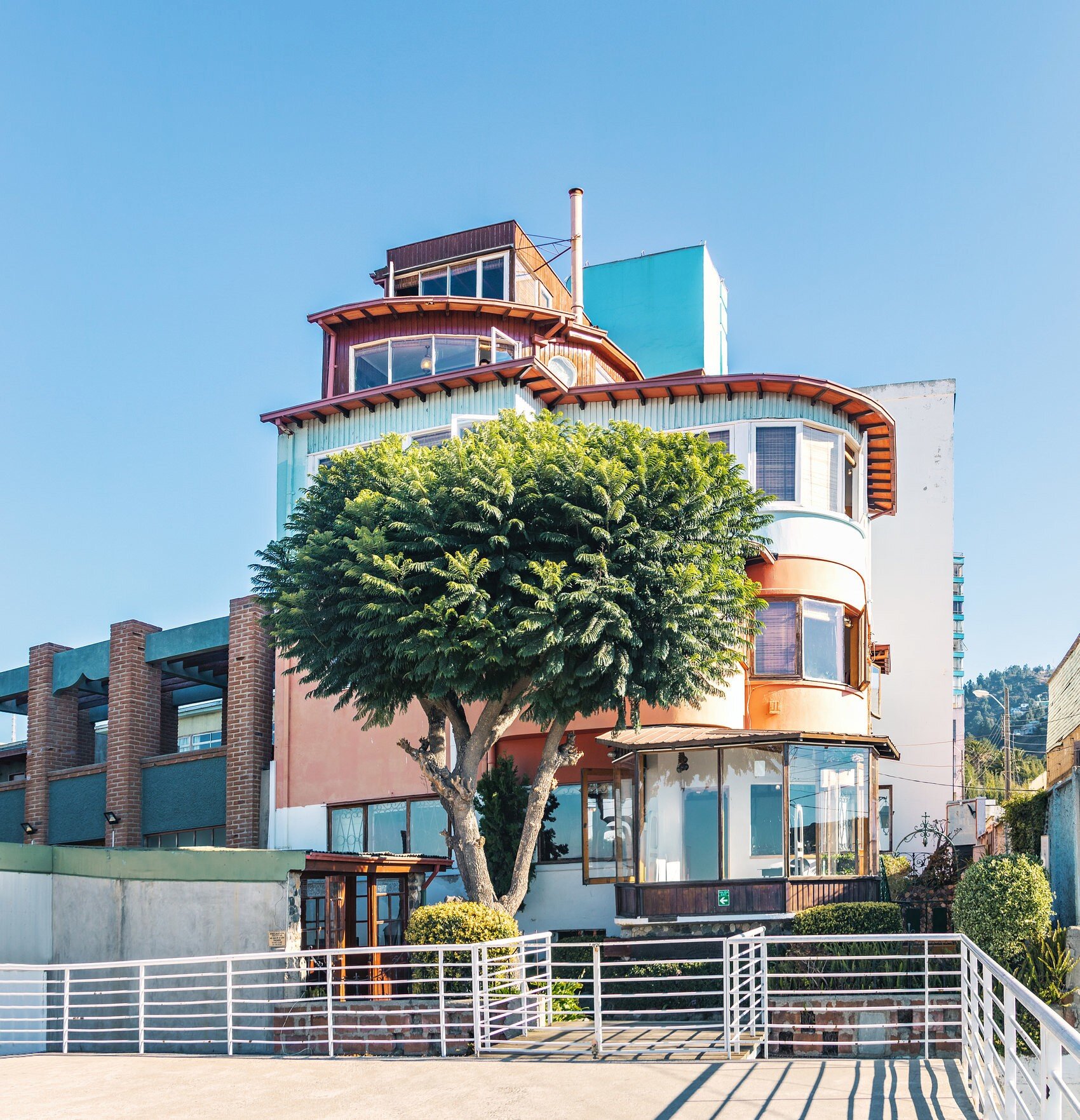 Tiered house with curved balconies and wooden upper rooms behind a leafy tree and white railings under a blue sky