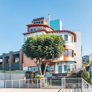 Tiered house with curved balconies and wooden upper rooms behind a leafy tree and white railings under a blue sky