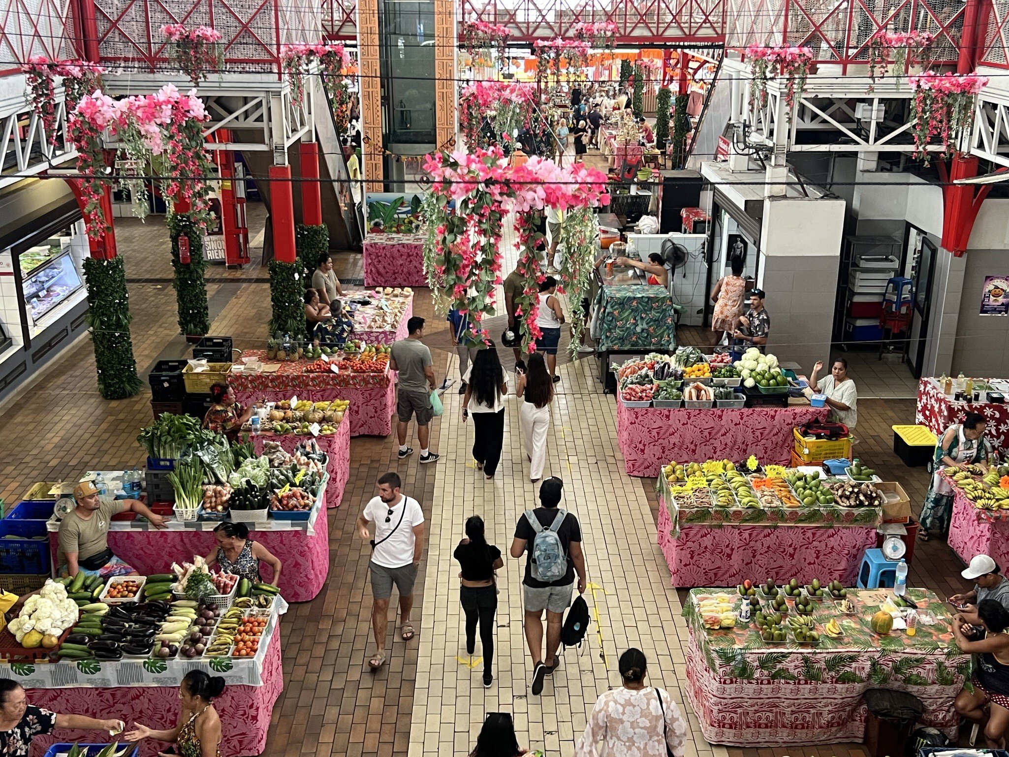 Market hall seen from above, fruit and vegetable tables draped in pink floral cloth, shoppers in the aisle.